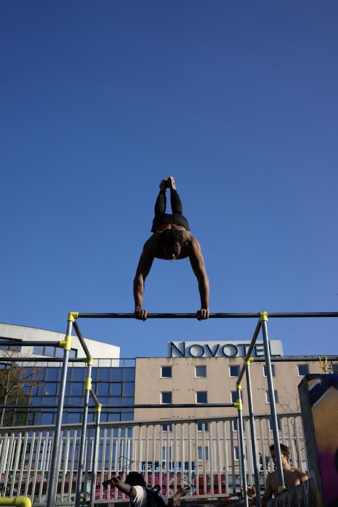 Athlète en figure sur une barre de street workout, ciel bleu et bâtiment urbain en arrière-plan.
